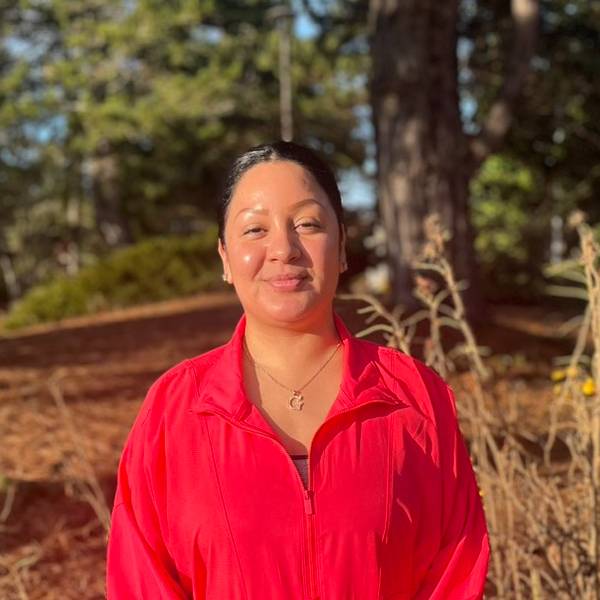 A headshot of Griselda. She smiles against a wooded background, wearing a bright red jacket.