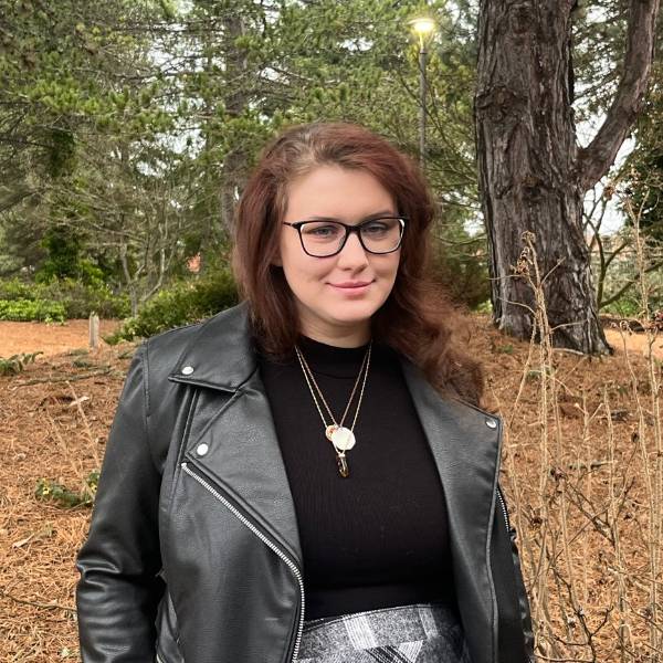 A headshot of Taylor. She smiles against a wooded background, wearing a black shirt and black leather jacket.