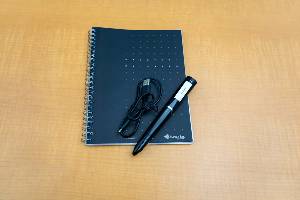 large black digital pen with charging cable and notebook sitting on desk