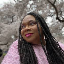 A headshot of an artist wearing a pink sweater surrounded by blossoming trees