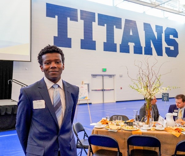 A student in a suit smiles in front of a wall that says "Titan"