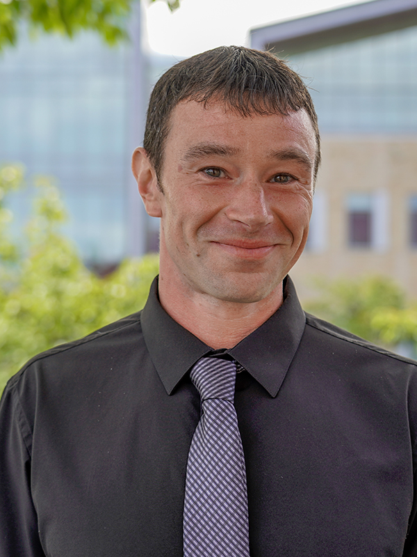 Picture of a man in a black shirt with a blue tie smiling. 